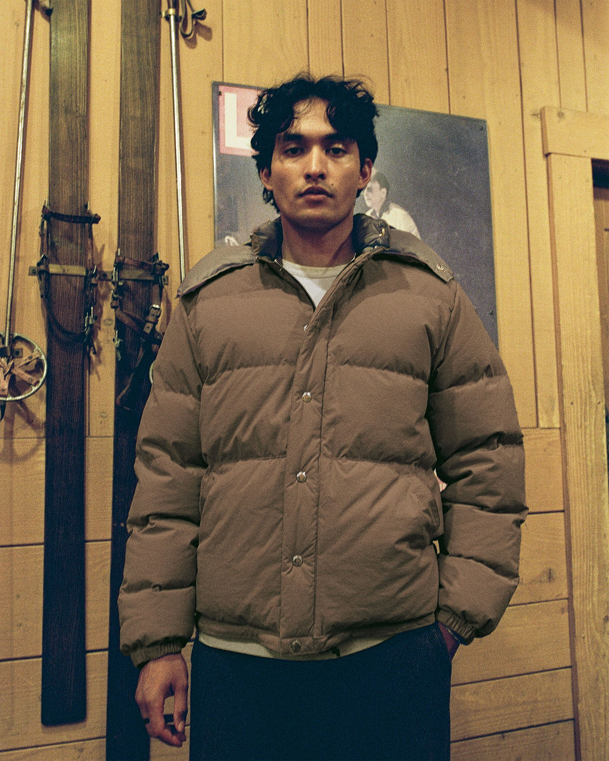 Man wearing a brown puffer jacket standing in a room with wooden walls and skis.
