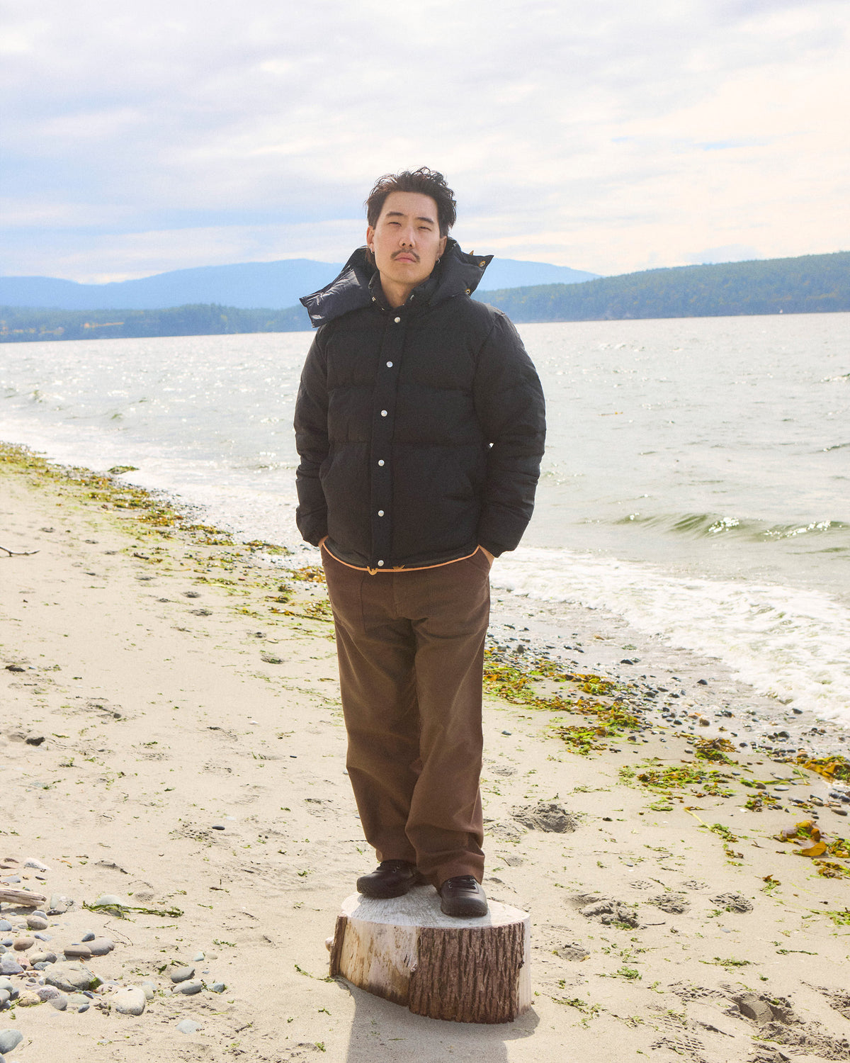 Man wearing black down sweater with dry wax cotton hood and brown pants standing on tree stump by beach