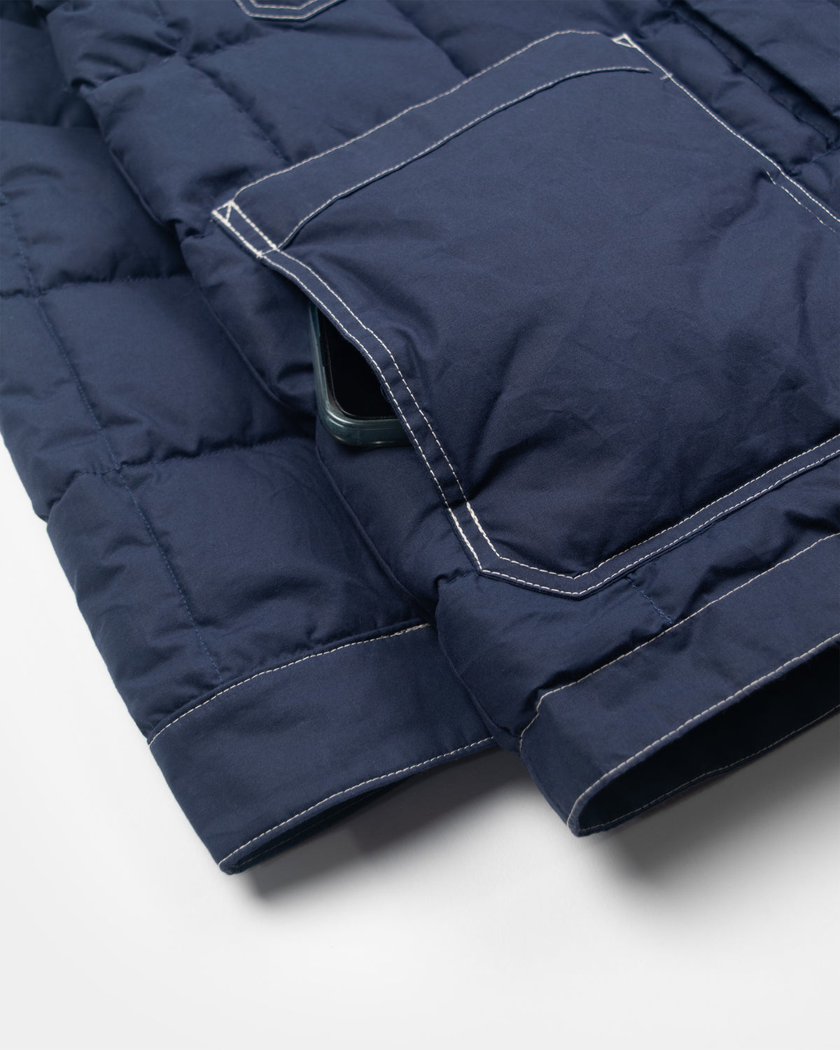 Close-up of a navy blue down chore coat puffer jacket showing a pocket with cream contrast stitching and a dark green phone tucked into the pocket, on a light gray background.