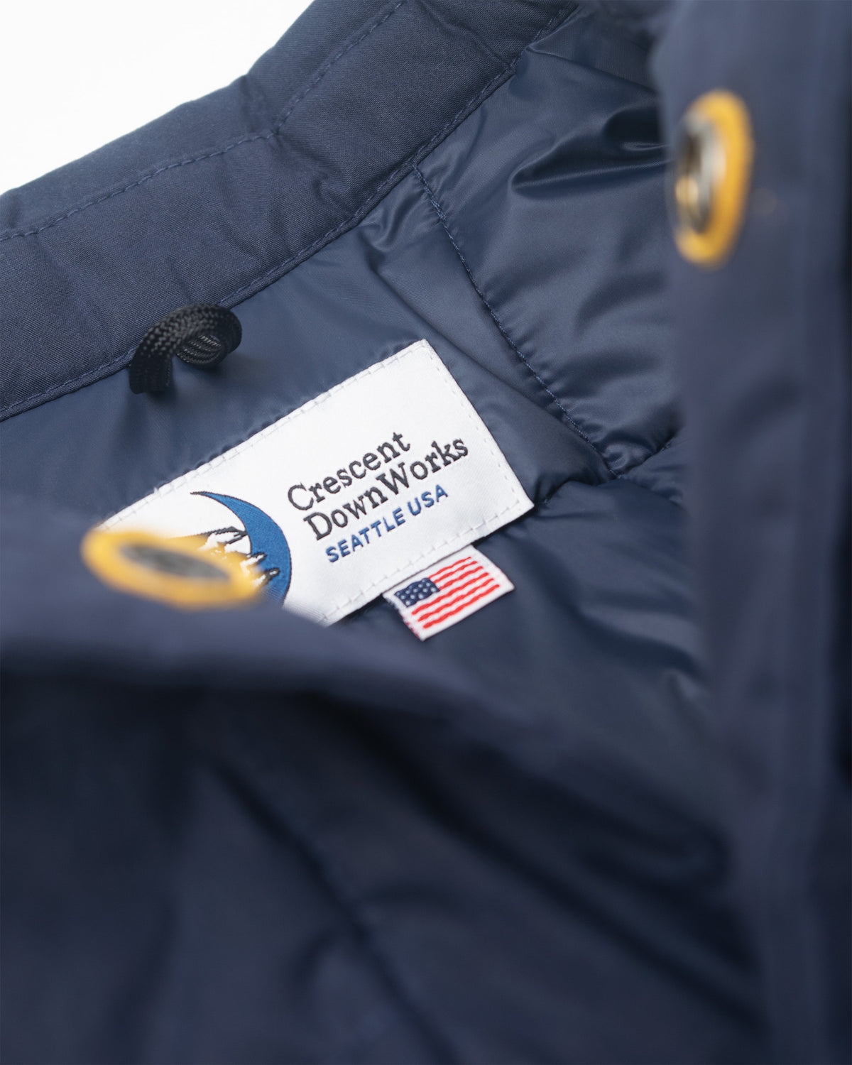 Close-up of a navy blue down chore coat jacket interior showing a white sewn-in label that reads 'Crescent Down Works SEATTLE USA' with a blue crescent logo, plus a small American flag tag.