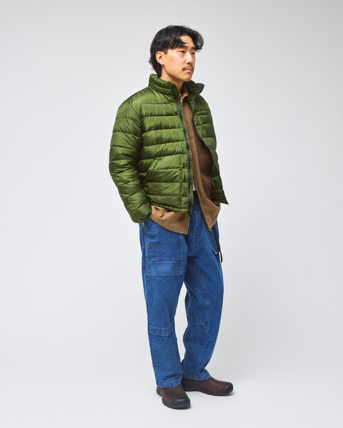 A man wearing an green Seattle Liner jacket over a brown shirt, blue jeans, and brown shoes, standing against a light gray studio background.