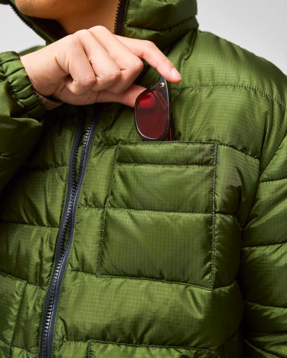 A model pulling sunglasses out of a chest pocket and wearing a green Seattle Liner jacket with horizontal quilting, against a light gray background.