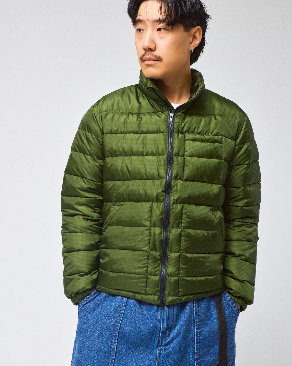 A model wearing a green Seattle Liner jacket with horizontal quilting, against a light gray background.