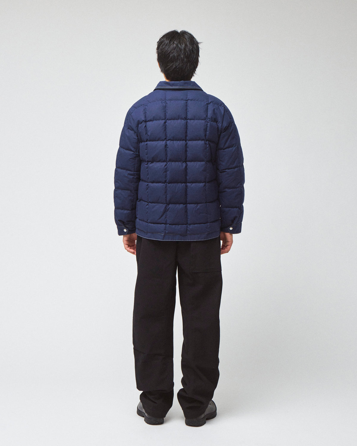 A man viewed from behind wearing a navy blue down chore coat puffer jacket, black pants, and black shoes, standing against a light gray studio background.
