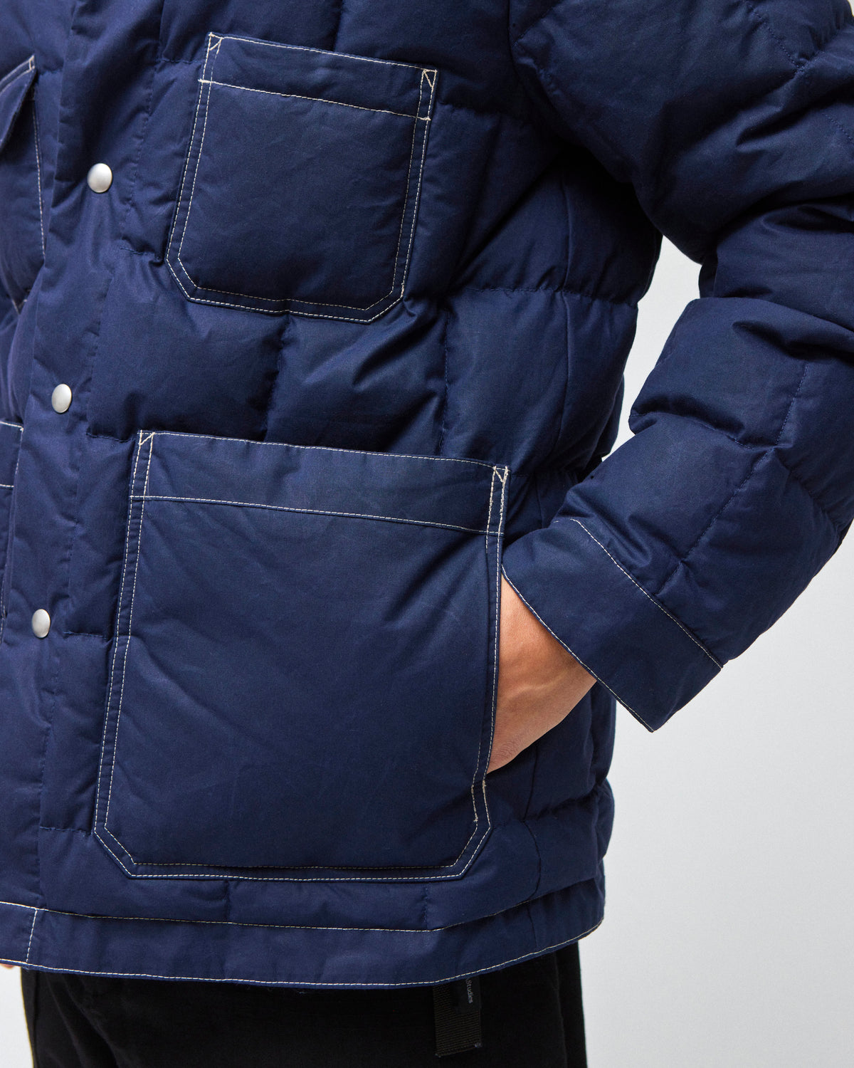 Close-up of a navy blue quilted down chore coat jacket with large front flap pockets and silver snap buttons, worn by a model whose hand is in a pocket, against a light gray background.