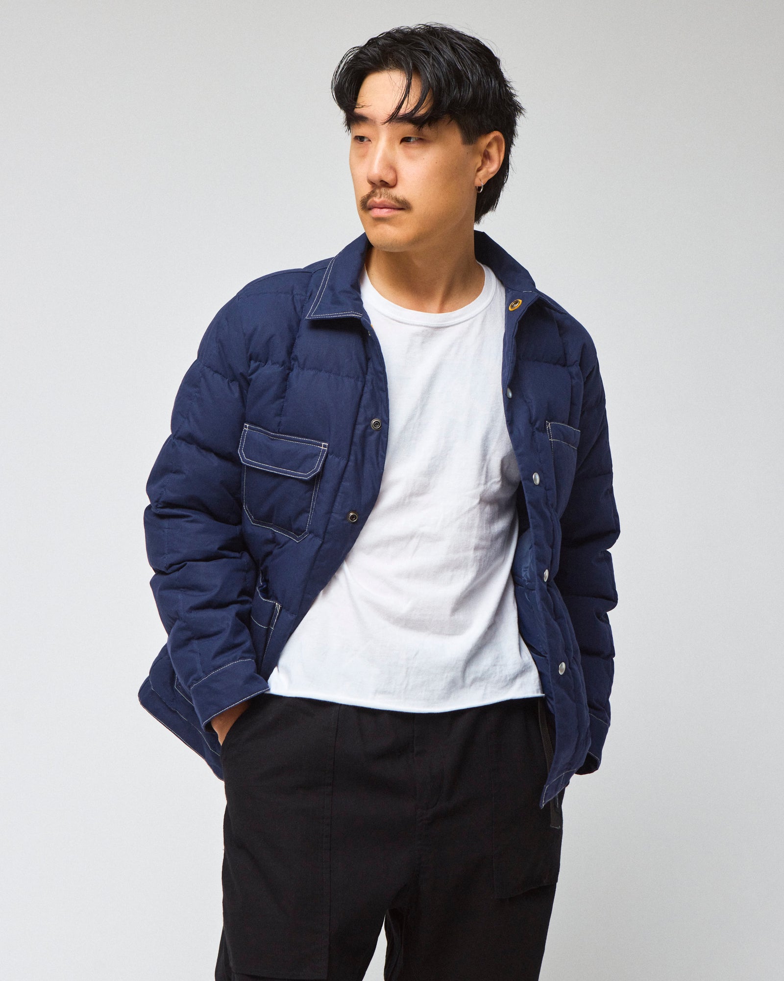 A man wearing a navy blue down chore coat jacket over a white t-shirt and black pants, standing against a light gray studio background.