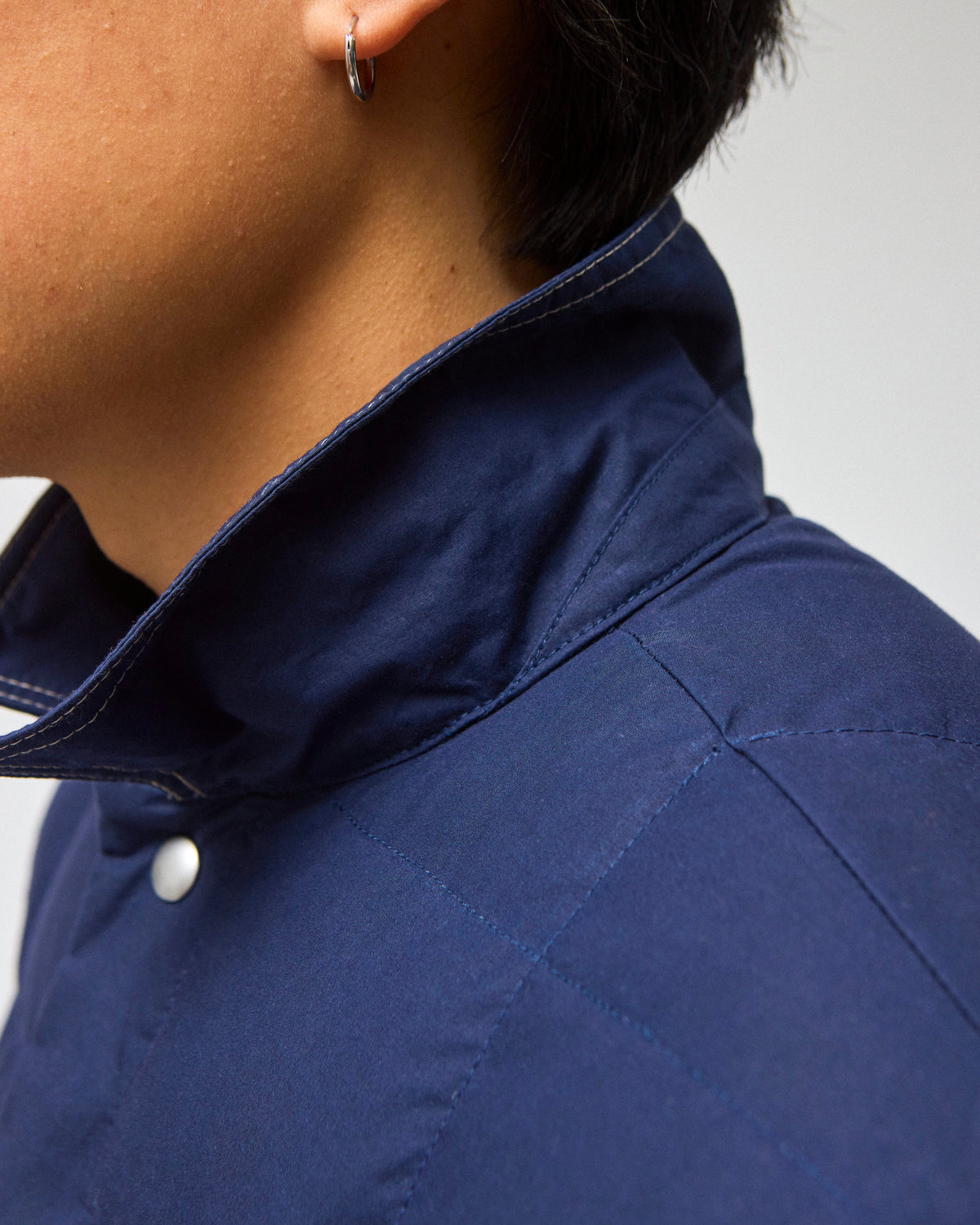 Close-up of a person wearing a navy blue quilted down chore coat jacket with a high collar and a silver hoop earring, against a light gray background.