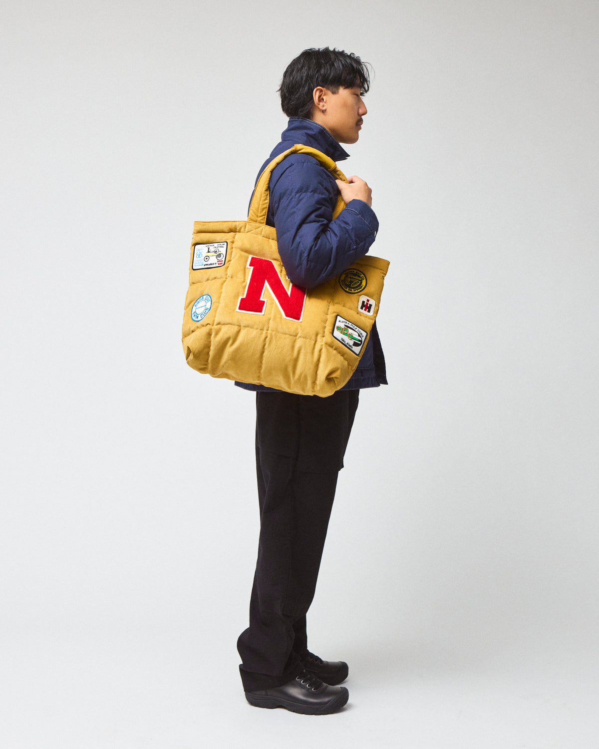 Model wearing a navy chore coat and black pants is shown from the side, carrying a yellow corduroy quilted tote bag with a large red letter 'N' on the front and several colorful patches.