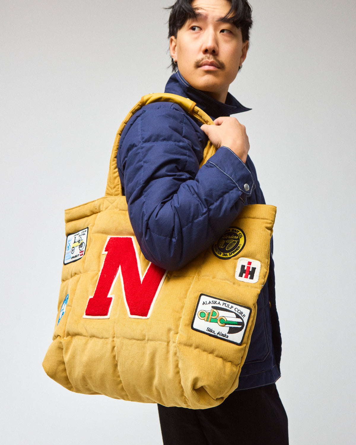 Model wearing a navy chore coat and black pants is shown carrying a yellow corduroy quilted tote bag with a large red letter 'N' on the front and several colorful patches.