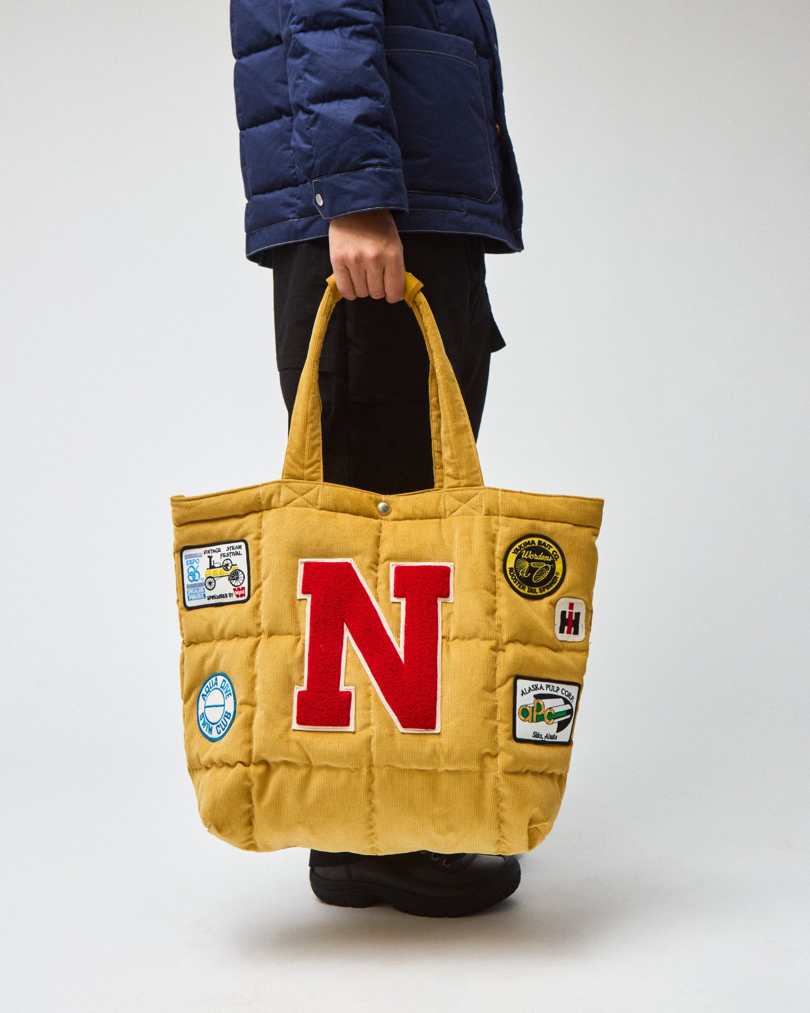 Model wearing a navy chore coat and black pants is shown from the side, carrying a yellow corduroy quilted tote bag with a large red letter 'N' on the front and several colorful patches.