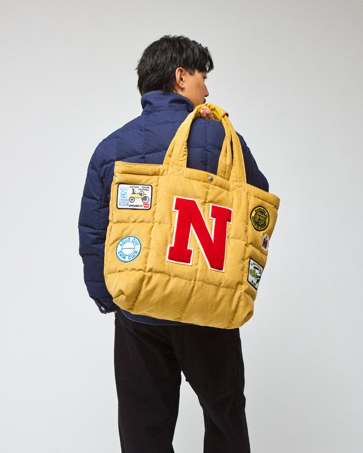 Model wearing a navy chore coat and black pants is shown from the back, carrying a yellow corduroy quilted tote bag with a large red letter 'N' on the front and several colorful patches.