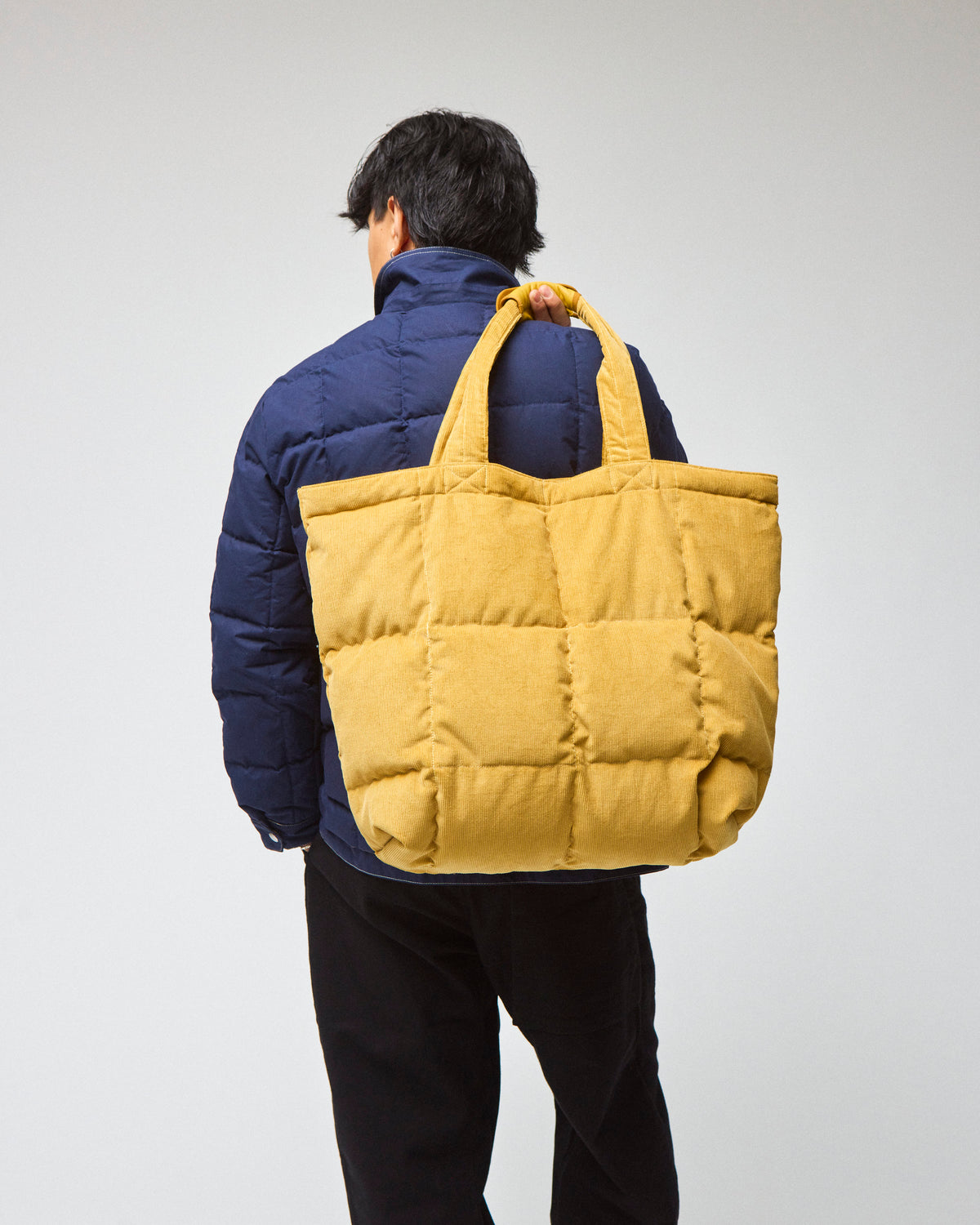 Model wearing a navy chore coat and black pants is shown from the back, carrying a yellow corduroy quilted tote bag over his shoulder.