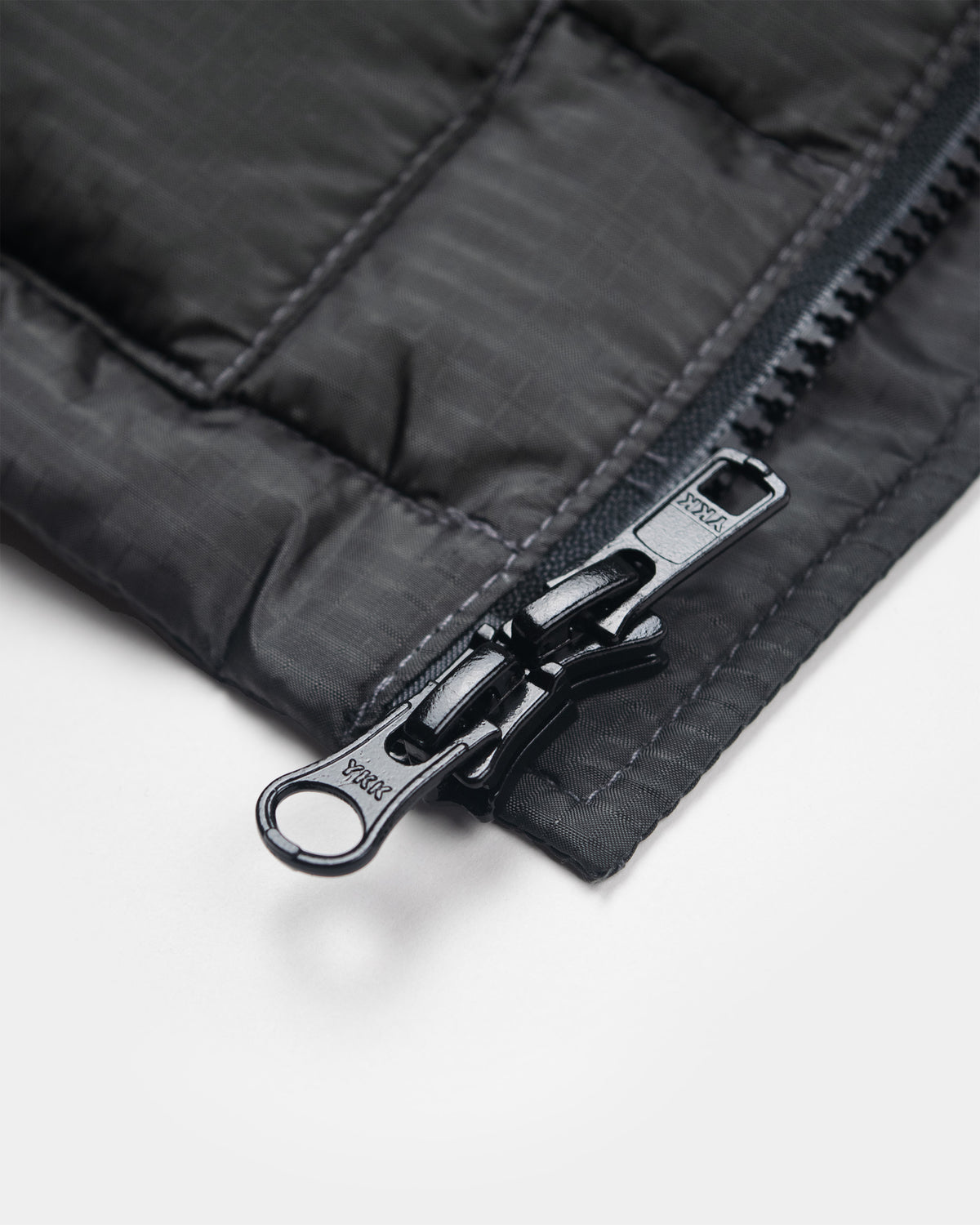 Close-up of a charcoal liner jacket corner showing a black metal zipper with a keyring pull tab on a white background.
