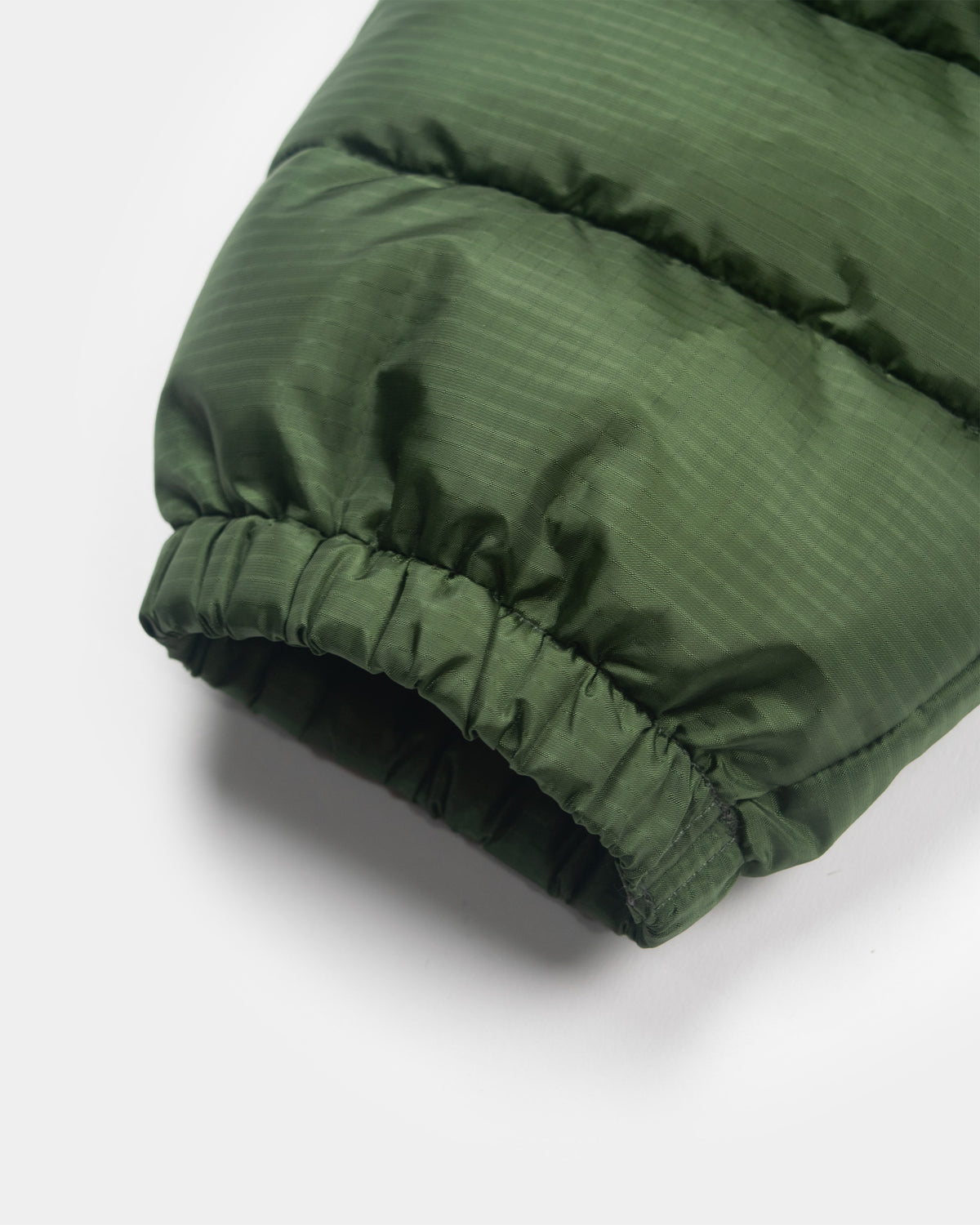 A detail closeup of the cuff of a green Seattle Liner jacket with horizontal quilting, against a light gray background.