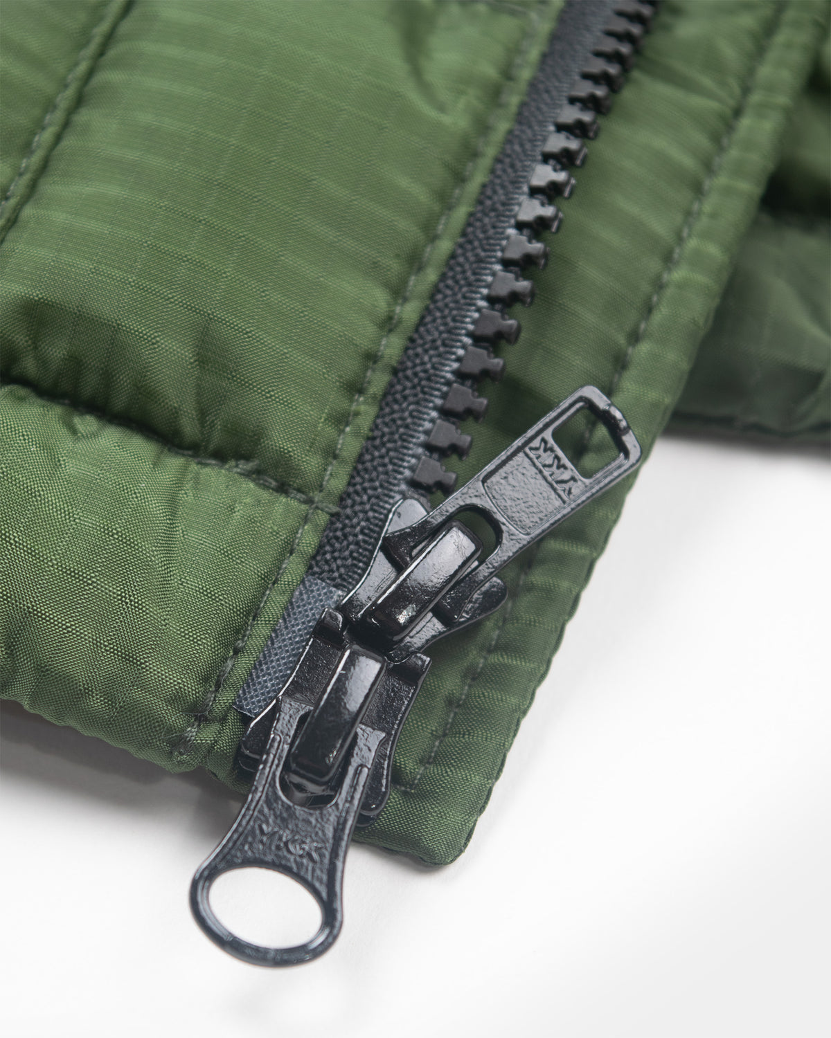 A detail closeup of the ykk zipper of a green Seattle Liner jacket with horizontal quilting, against a light gray background.