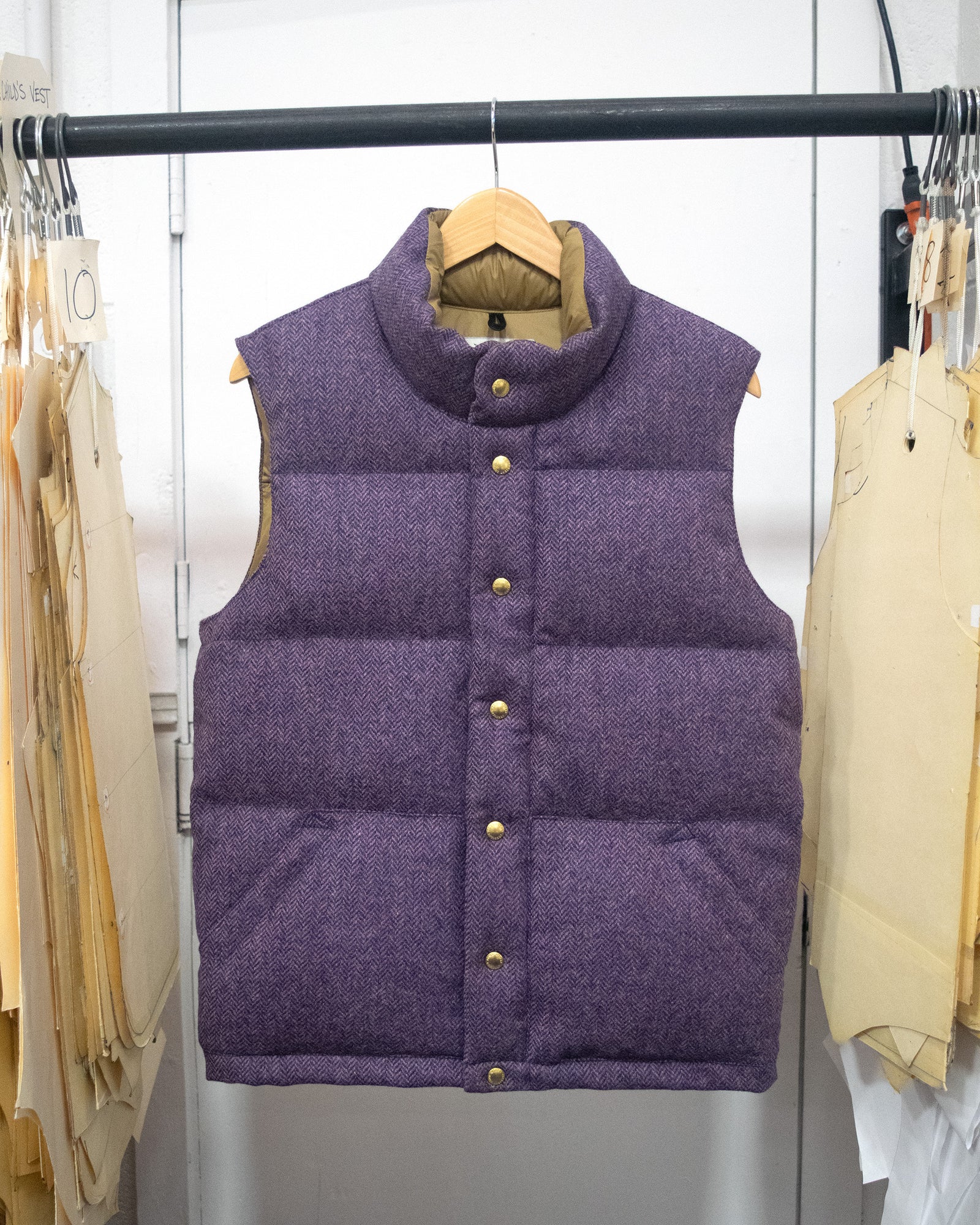 A purple quilted puffer vest with  brass tone snap buttons, hanging on a wooden hanger on a clothing rack.