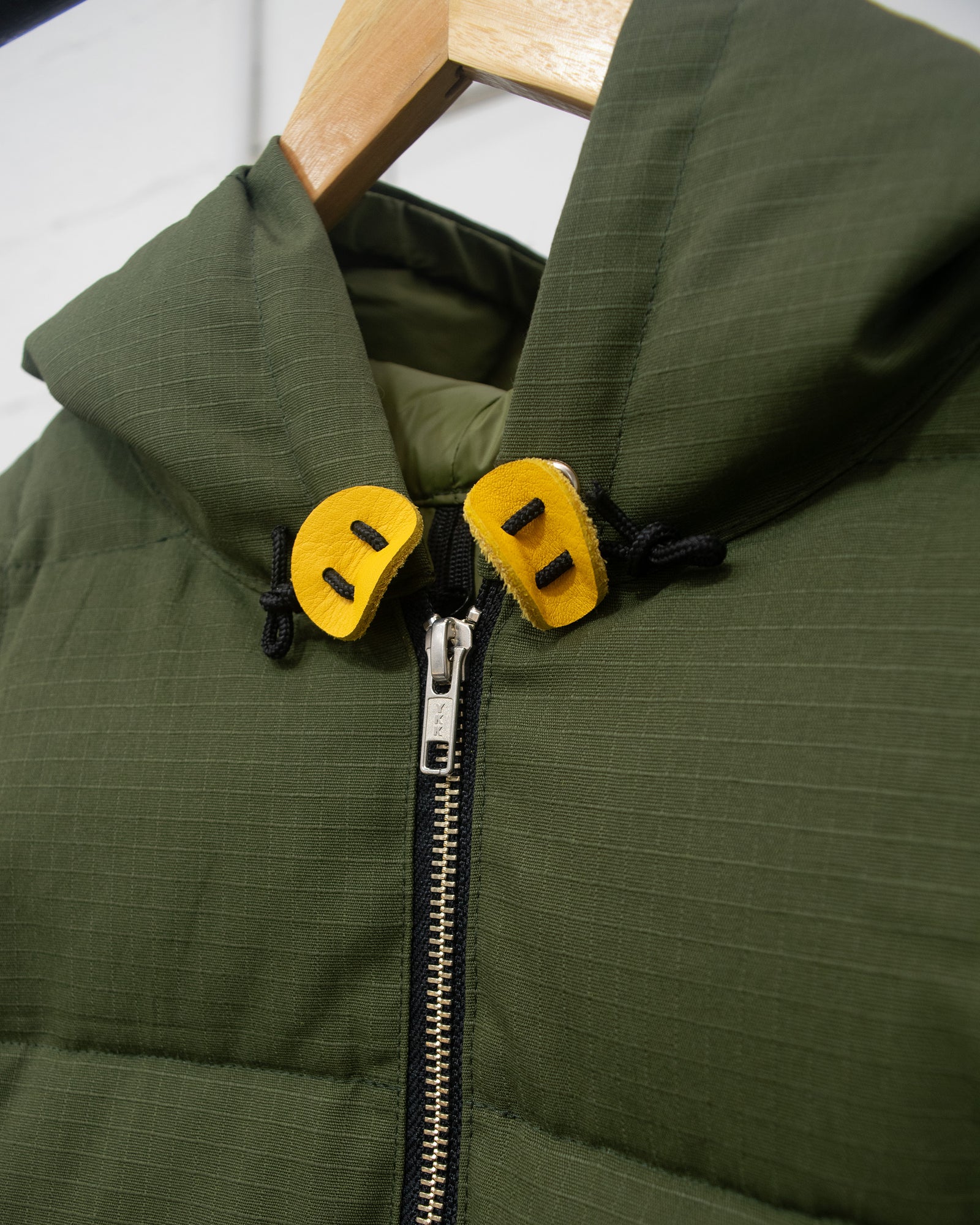 An olive green hooded sweatshirt puffer jacket with a black front zipper and yellow toggle details, hanging on a wooden hanger on a clothing rack.