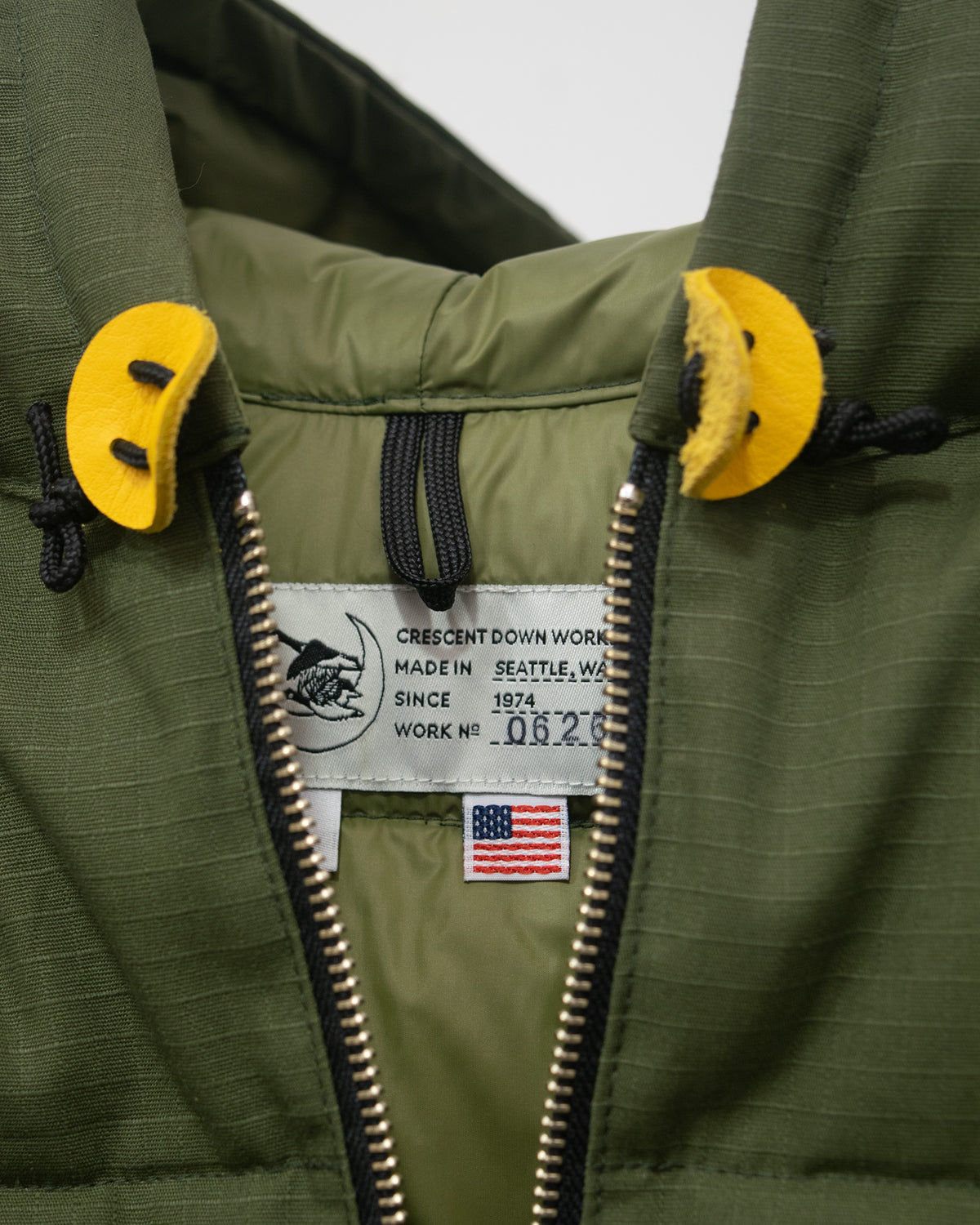 Close-up of an olive green hooded jacket with a black zipper, black drawstrings, and bright yellow leather toggles. The inner label reads Crescent Down Work, Made in Seattle, WA, and includes a small American flag patch.