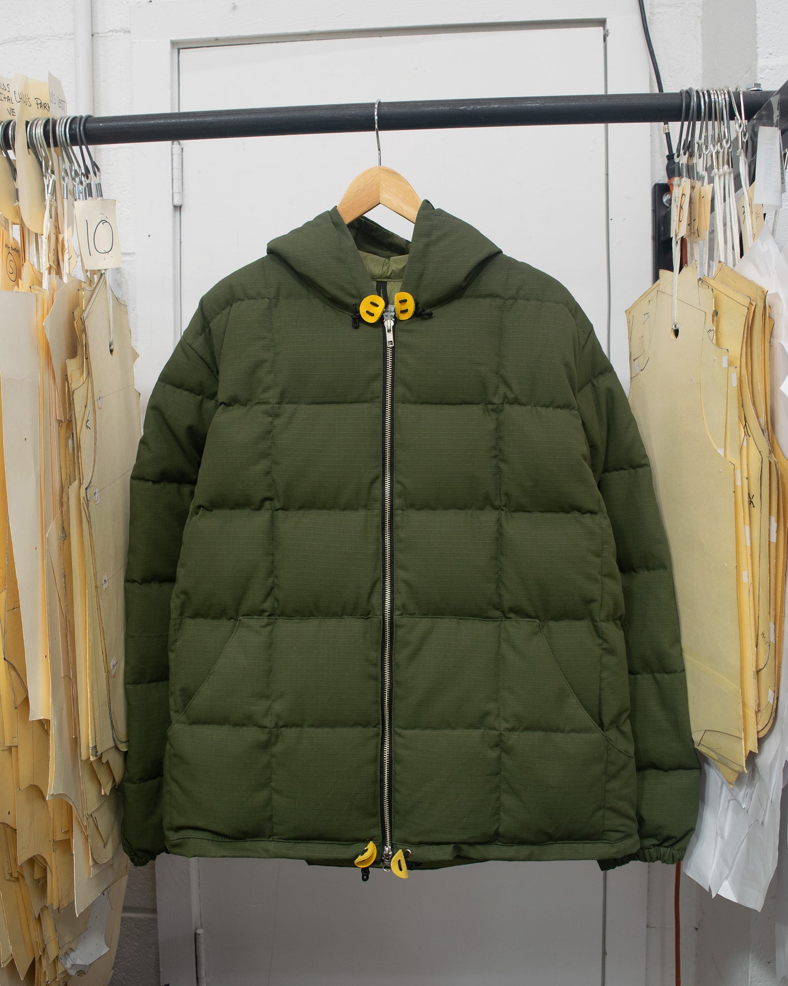 An olive green hooded sweatshirt puffer jacket with a black front zipper and yellow toggle details, hanging on a wooden hanger on a clothing rack.