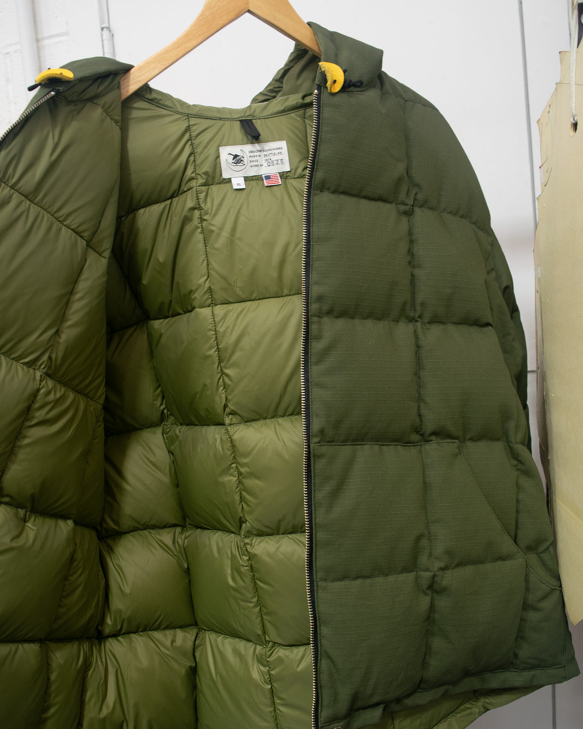 An olive green puffer jacket hanging on a wooden hanger against a white wall, showing the front zipper, quilted padding, and a small white brand label.