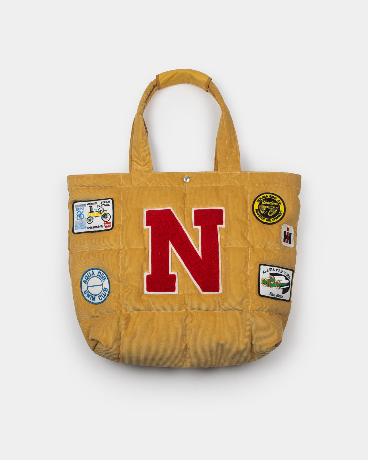 A yellow corduroy tote bag featuring a large red block letter 'N' outlined in white on the front, decorated with several colorful vintage-style patch designs on the sides, set against a light gray background.