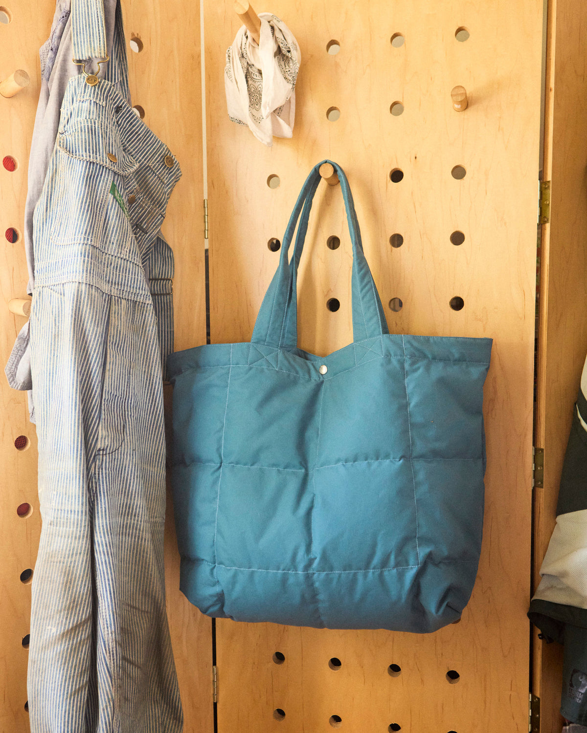A Robin blue puffer tote bag hanging on a wooden pegboard, next to a striped shirt and other hanging items.