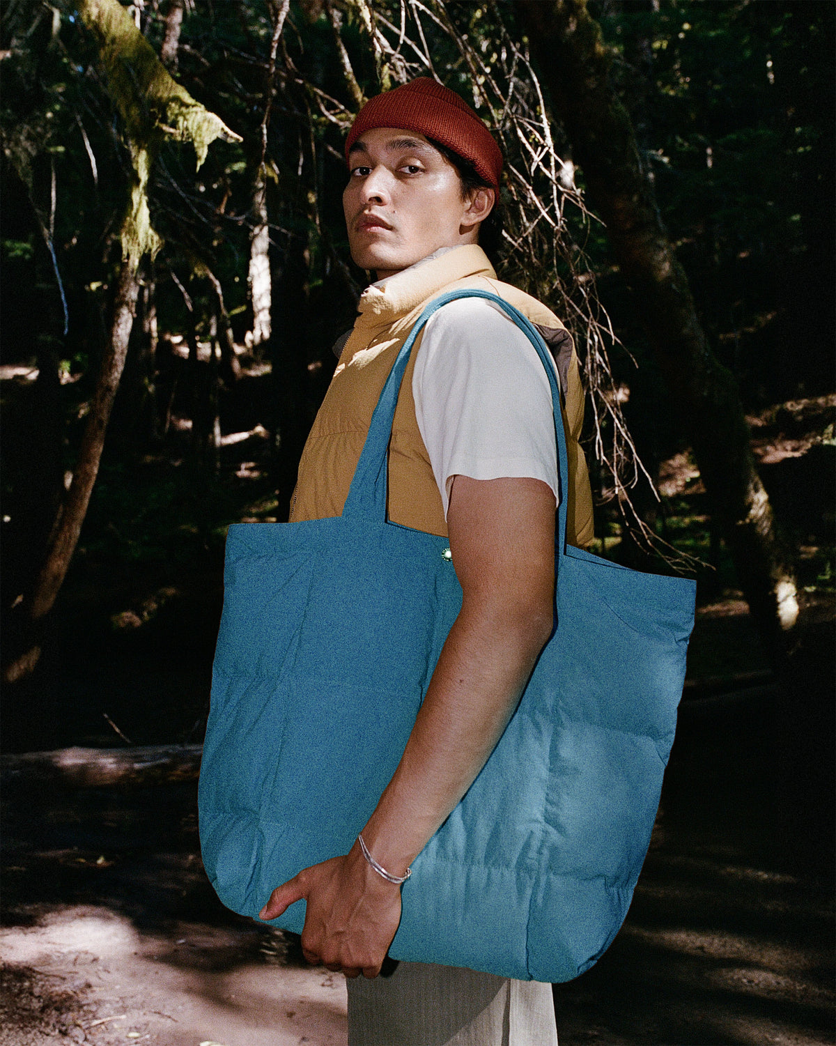 A person standing in a forest holding a large Robin Blue puffer tote bag. The person is wearing a red beanie, a tan North by Northwest vest over a white t-shirt, and light-colored pants.