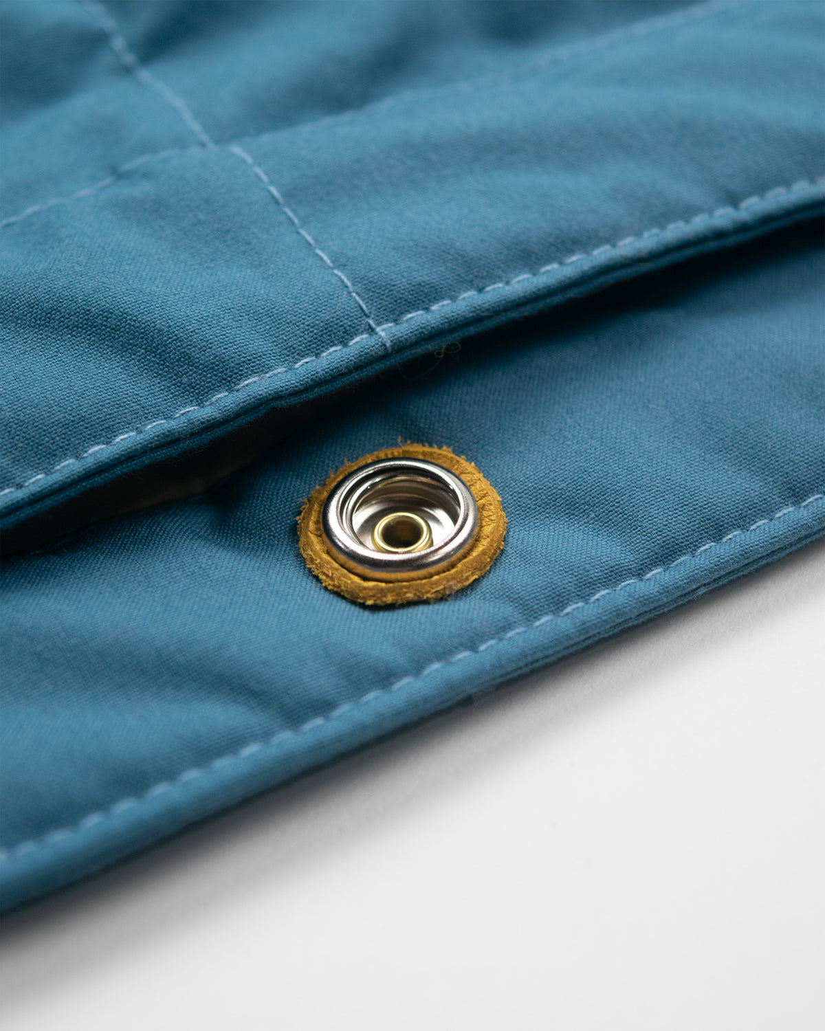 Close-up of a Robin blue dry wax cotton quilted fabric with snap button closure reinforced with gold leather on a white background.