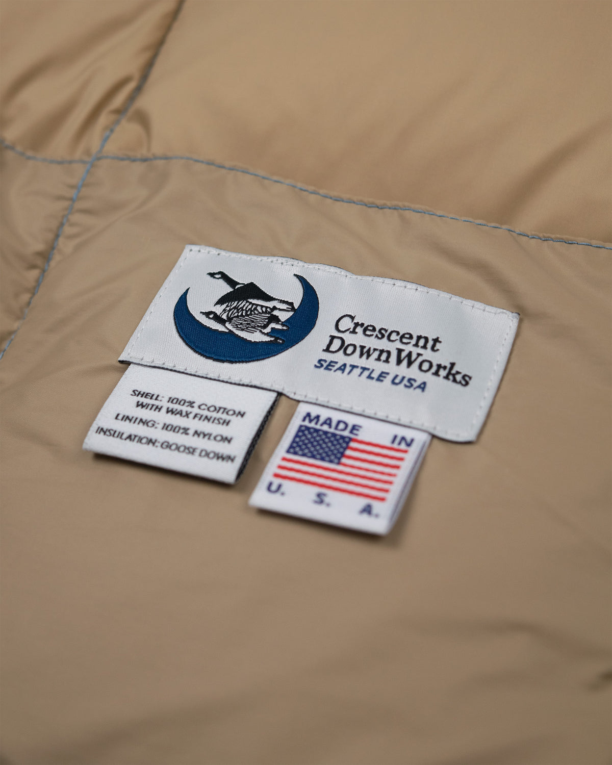 Close-up of an amber quilted down tote bag showing two sewn-in labels: a white Crescent Down Works Seattle USA label with a blue logo, and a smaller white Made in U.S.